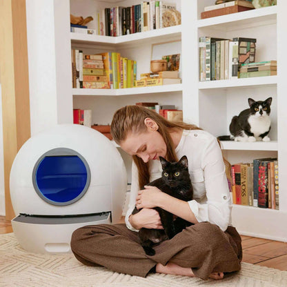 Woman with a black cat beside a smart self-cleaning litter box in a cozy home setting.